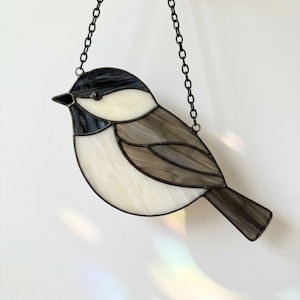 May include: A stained glass suncatcher shaped like a chickadee bird, featuring a black head, white cheeks, and grey wings and tail. The bird is constructed with dark metal solder lines and is suspended by a thin black metal chain for hanging.