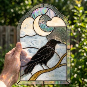 Op de afbeelding: Een glas-in-loodpaneel met een zwarte kraai op een tak tegen een achtergrond van witte wolken en een maansikkel. Het werk is opgebouwd met donkere loden lijnen en bevat iriserende glasaccenten in de maan en de lucht.