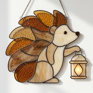 May include: A stained glass suncatcher shaped like a hedgehog with a cream-colored body and textured amber-brown spines. The hedgehog holds a small lantern with a glowing yellow light. The piece is framed in dark metal and hangs by a thin chain.