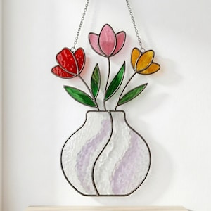 May include: A stained glass suncatcher featuring a vase with three tulips in red, pink, and yellow. The vase is made of textured white glass with purple accents. The piece is suspended by a metal chain, showcasing green glass leaves and stems.