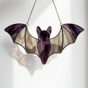 Puede incluir: Un atrapasoles de vidriera con forma de murciélago, compuesto por paneles de vidrio morado oscuro y blanco translúcido unidos por líneas de soldadura de metal oscuro. El murciélago cuelga de una fina cadena metálica sujeta a las alas.
