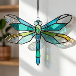 May include: Stained glass dragonfly with a black metal frame. The dragonfly features iridescent wings in shades of blue, green, and yellow. The dragonfly is hanging against a white wall, with a black string attached to the top.
