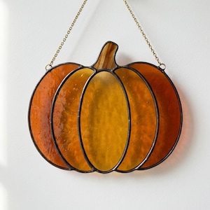 May include: Stained glass pumpkin decoration with a black outline and a gold chain for hanging. The pumpkin features shades of orange and yellow glass, creating a warm, autumnal aesthetic. A decorative piece for autumn.