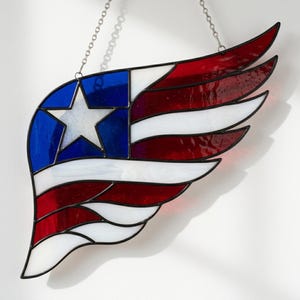 May include: A stained glass wing in the colors of the American flag, featuring a blue section with a white star and red and white stripes. The wing is outlined in black and hangs from a silver chain.