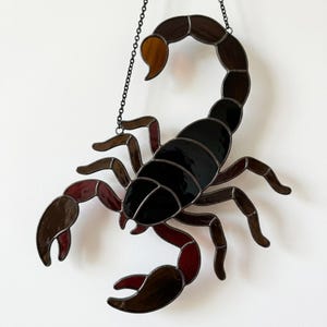May include: A stained glass scorpion suncatcher featuring black, dark brown, and amber glass segments held together by dark metal solder lines. The scorpion is suspended by a thin black metal chain attached to its curved tail and body.