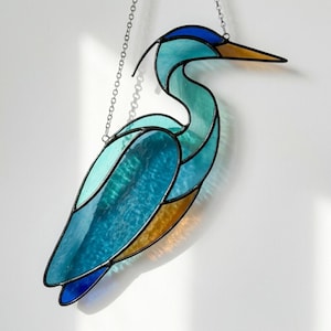 May include: Stained glass heron in shades of blue, teal, and gold, hanging from a silver chain. The bird's form is outlined in black, with a long beak and elegant neck. A decorative piece for windows or walls.