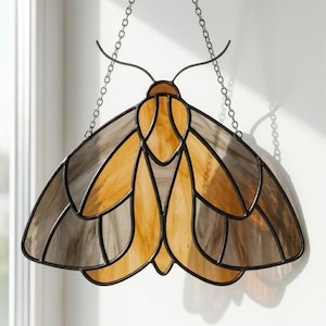 May include: Stained glass moth suncatcher with a black metal frame. The wings are a mix of amber and gray, creating a gradient effect. The suncatcher hangs from a chain, adding a decorative touch to any window or wall.