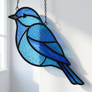 May include: A stained glass bird decoration in shades of blue, with a black outline and a chain for hanging. The bird features textured glass and a detailed design, perfect for adding a touch of nature-inspired art to any space.
