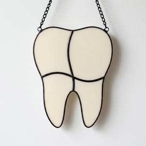 May include: A stained glass hanging ornament shaped like a human tooth, featuring off-white glass panels held together by dark metal solder lines. The piece is suspended by a thin black metal chain attached to the top of the tooth-shaped frame.