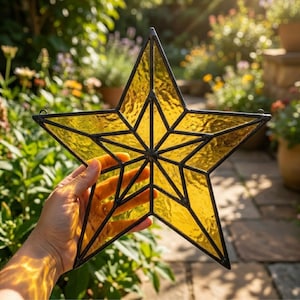 May include: A yellow stained glass star with a black metal frame. The star has a textured surface and is held up in front of a garden background. The star is a decorative item.