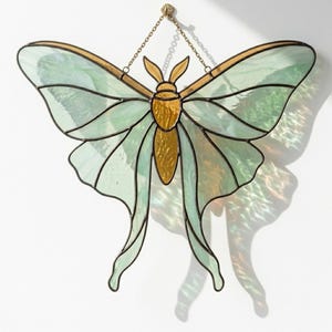 Pode incluir: Um apanhador de sol de vitral em forma de traça com asas verde-menta pálido e linhas de soldadura de metal escuro. O corpo central e as antenas são feitos de vidro texturizado cor de âmbar. Uma fina corrente de metal está presa no topo.