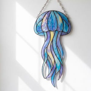 May include: Stained glass jellyfish wall hanging in shades of blue, purple, and yellow. The jellyfish has a rounded bell and long, flowing tentacles. It is suspended by a silver chain, creating a decorative ocean-themed art piece.
