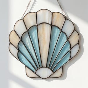 May include: Stained glass seashell wall hanging with a chain. The shell design features white, light blue, and beige glass panels, framed with dark metal. A decorative coastal accent for the home.