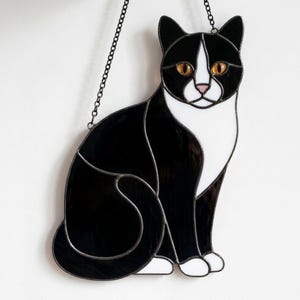 Op de afbeelding: Een glas-in-lood suncatcher in de vorm van een zittende tuxedo kat met zwarte en witte glazen panelen, amberkleurige ogen en een roze neus. De katfiguur is omlijnd met donker metaalsoldeer en hangt aan een dunne zwarte metalen ketting.