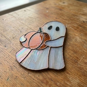 May include: A stained glass suncatcher shaped like a ghost holding an orange pumpkin. The ghost is made of iridescent white glass with two black glass eyes, while the pumpkin features textured amber glass. The pieces are joined by dark metal solder.