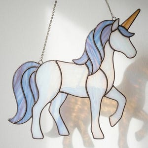 Puede incluir: Un atrapasoles de vidriera con forma de unicornio, cuerpo blanco iridiscente y una crin y cola fluidas en tonos azul claro y morado. El unicornio tiene un cuerno amarillo dorado y está suspendido mediante una fina cadena de metal.