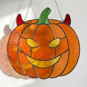 Può includere: Un acchiappasole in vetro colorato a forma di zucca con vetro arancione testurizzato, un picciolo verde, occhi e bocca gialli e due piccole corna da diavolo rosse. L'oggetto è realizzato con linee di saldatura in metallo scuro e una catena.