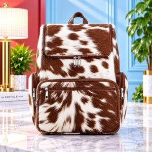 May include: A backpack featuring a brown and white cow print pattern with a textured hair-on-hide appearance. The bag includes a top handle, a large main compartment with a zippered flap, a front zippered pocket, and side zippered pockets.