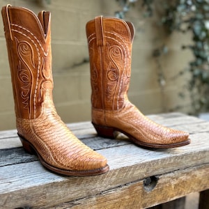 May include: A pair of brown cowboy boots with intricate stitching and a textured, snake-like pattern on the lower part. The boots are made of leather and have pointed toes and stacked heels. The boots are displayed on a wooden surface.
