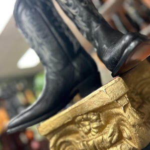 Könnte beinhalten: Ein Paar schwarze Leder-Cowboystiefel mit strukturierter Oberfläche am Fuß und aufwendigen Ziernähten am hohen Schaft. Die Stiefel haben eine spitz zulaufende Kappe, einen Blockabsatz und eine glatte Sohle und stehen auf einem goldenen Sockel.