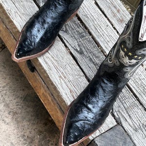 May include: A pair of black cowboy boots with pointed toes and decorative stitching. The boots have a textured leather appearance and a brown sole. The upper part of the boots features intricate silver and gold embroidery.