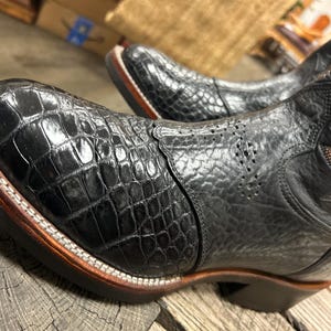 Könnte beinhalten: Ein schwarzer Westernstiefel mit einer glänzenden, texturierten Zehenkappe in Krokodilleder-Optik und einem matten Schaft aus genarbtem Leder. Der Stiefel verfügt über einen braunen Absatz, weiße Rahmennähte und dekorative braune Stickereien.