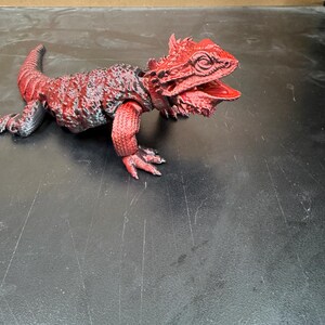 Op de afbeelding: Een rood en zwart gearticuleerd hagedisfiguur met een getextureerd, geschubd oppervlak en een open mond. De hagedis heeft een stekelige kop, een lange staart en vier poten met zichtbare klauwen, met een gedetailleerde, meerkleurige afwerking.