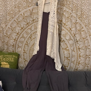 May include: A sleeveless cream-colored lace vest hangs over a dark purple jumpsuit. The jumpsuit has a wide leg and gathered ankles. The outfit is displayed on a dark gray couch in front of a patterned tapestry.