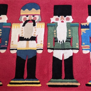 May include: A red rug with four nutcracker figures. The nutcrackers are wearing colorful uniforms and have white beards. The rug is made of a soft, plush material.