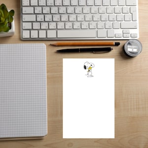 May include: A white sheet of paper with a Snoopy illustration holding a yellow flower. A pencil, pen, and keyboard are visible on a wooden desk. A notebook and a small plant are also in the image.
