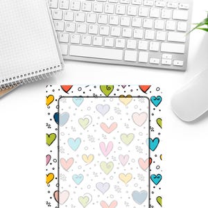 Pode incluir: Um tapete de secretária com um padrão de corações coloridos, redemoinhos e pontos pretos sobre um fundo branco. Um bloco de notas branco e um teclado de computador estão posicionados acima do tapete. Os corações são de várias cores.