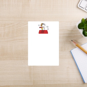 May include: A white notepad with a cartoon illustration of Snoopy wearing a scarf and goggles on top of his red doghouse. The image is on a light wood-grain surface with a pencil, keyboard, and plant in the background.