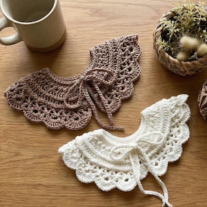 May include: Two crocheted collars, one in light brown and the other in white, are shown on a wooden surface. Each collar has a scalloped edge and a tie closure. The collars are handmade with a delicate, openwork design.