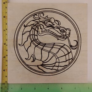 Op de afbeelding: Een houten vierkant met een zwart drakenontwerp dat in het oppervlak is geëtst. De draak staat in een cirkelvormig frame, met zijn kop en nek zichtbaar. Het ontwerp is ongeveer 10 cm vierkant.