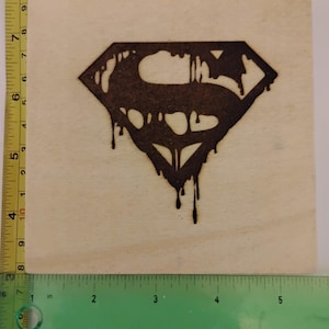 May include: A wooden square with a burnt Superman logo design. The logo is a dark brown color and appears to be dripping. The wooden square measures approximately 4 inches by 4 inches.