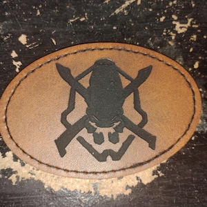 May include: Oval-shaped brown leather patch with a black skull and crossed swords design. The patch is stitched around the edge with brown thread. The design is a military or tactical emblem.