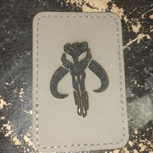 Puede incluir: Un parche rectangular con fondo gris y un emblema negro. El emblema es un diseño estilizado con formas curvas y líneas verticales. El parche tiene un borde cosido.