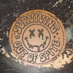 May include: A round, brown leather patch with the words "SURVIVING OUT OF SPITE" in black lettering. The patch features a smiley face with X eyes and dripping mouth. A butterfly and stars are also included.
