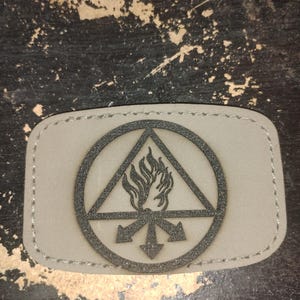 Op de afbeelding: Een rechthoekige patch met een lichtgrijze achtergrond en een zwart grafisch ontwerp. Het ontwerp toont een driehoek in een cirkel, met vlammen en pijlen. De patch heeft een gestikte rand.