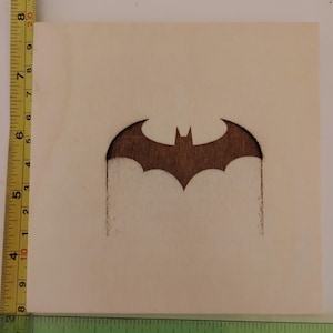 Puede incluir: Un cuadrado de madera con el logotipo de Batman marrón oscuro, una silueta de murciélago, quemado en el centro. La madera es de color claro. Una regla en la parte inferior muestra medidas en pulgadas, aproximadamente 10 cm.