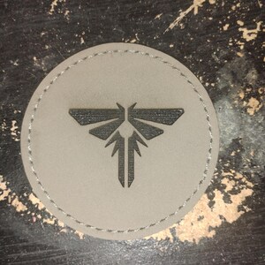 May include: A circular, gray patch with a stitched border. The patch features a black, stylized firefly emblem in the center. The design is a minimalist, geometric representation of the insect.