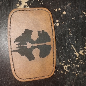 Puede incluir: Un parche de cuero marrón con un diseño de paisaje negro. El parche es rectangular con esquinas redondeadas y un borde cosido. El paisaje representa una silueta de montaña y su reflejo.