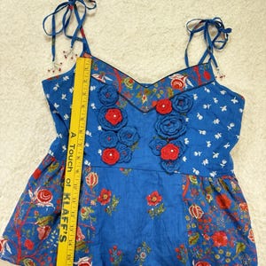 May include: Blue floral print top with red crochet flower accents. The top features adjustable tie straps and a V-neckline. The fabric has a mix of floral and bird patterns. The top is approximately 18 inches long.