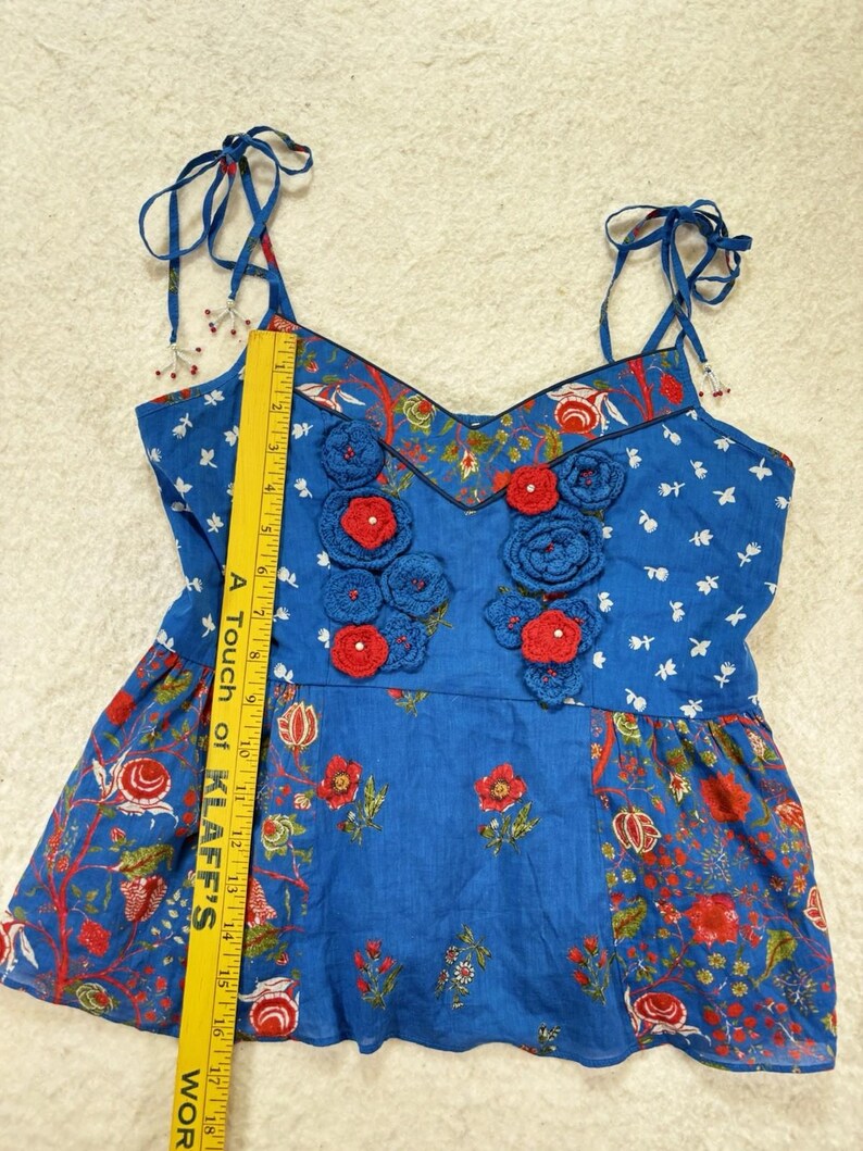 May include: Blue floral print top with red crochet flower accents. The top features adjustable tie straps and a V-neckline. The fabric has a mix of floral and bird patterns. The top is approximately 18 inches long.