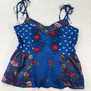 May include: A blue camisole top with floral and botanical patterns. The top features red crocheted flowers and tie straps. The neckline and straps are trimmed with a floral print. The bottom of the top has a ruffled hem.