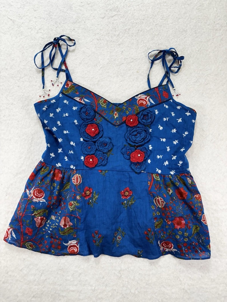 May include: A blue camisole top with floral and botanical patterns. The top features red crocheted flowers and tie straps. The neckline and straps are trimmed with a floral print. The bottom of the top has a ruffled hem.