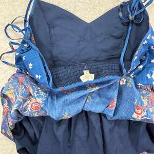 May include: A blue floral print sundress with adjustable tie straps. The dress features a V-neckline and a layered design. The fabric has a mix of red, white, and green floral patterns on a blue background. The dress has a small tag with the brand name.