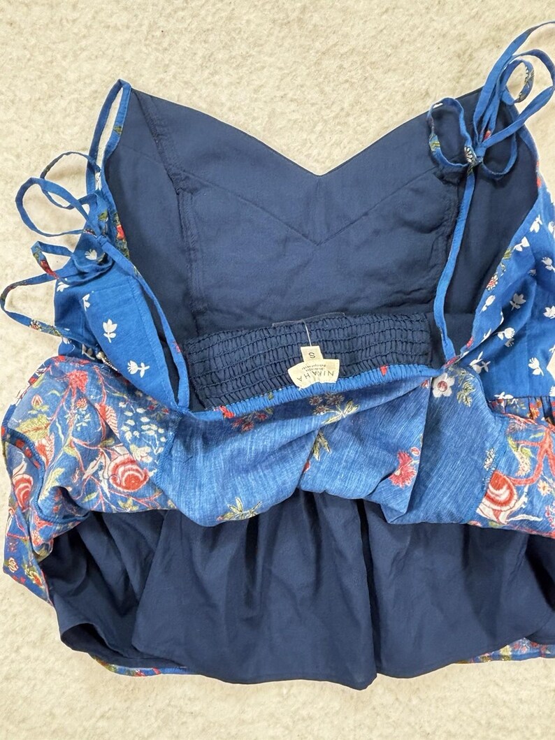 May include: A blue floral print sundress with adjustable tie straps. The dress features a V-neckline and a layered design. The fabric has a mix of red, white, and green floral patterns on a blue background. The dress has a small tag with the brand name.