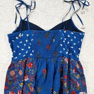 May include: A blue floral print top with adjustable tie straps. The top features a combination of patterns, including white floral accents and a red floral design. The bodice has a smocked detail, and the straps are adorned with small beads.