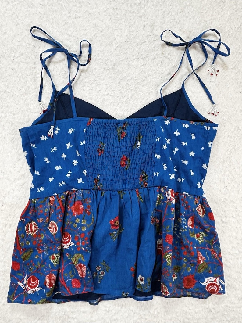 May include: A blue floral print top with adjustable tie straps. The top features a combination of patterns, including white floral accents and a red floral design. The bodice has a smocked detail, and the straps are adorned with small beads.